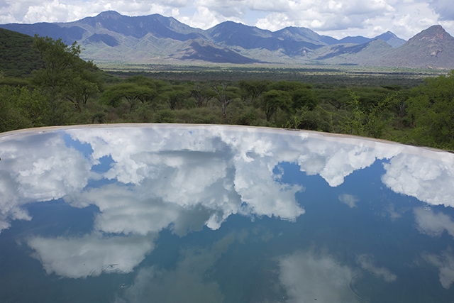 Image of clouds reflecting in water with trees and mountains in the background.