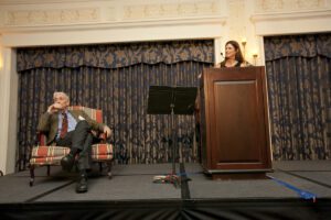 Image of a woman speaking on stage and E.O. Wilson sitting beside her.